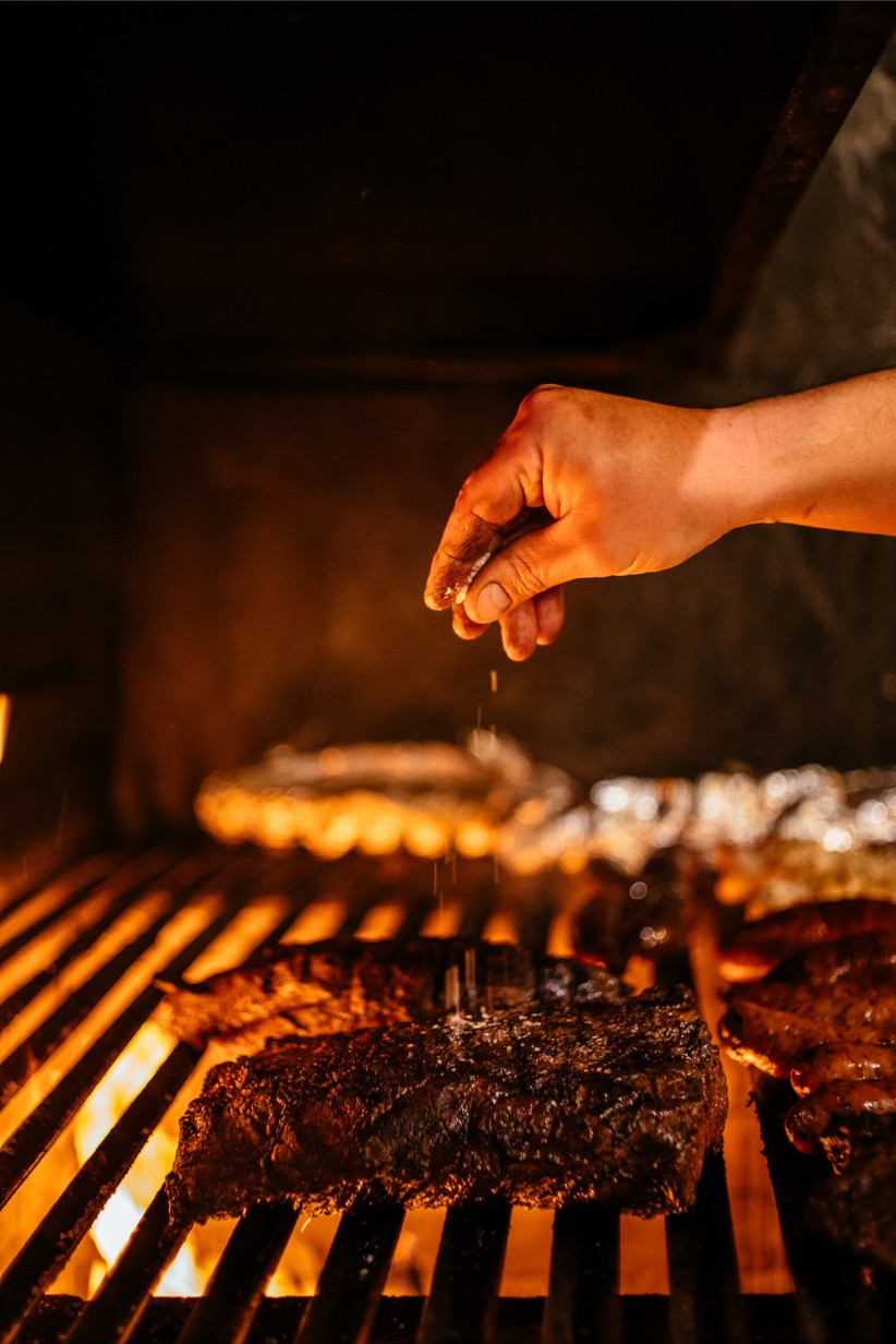 A chef's hand sprinkling salt on top of a large steak on the grill.