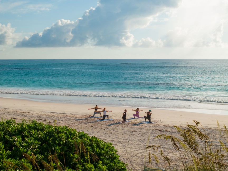 A group of people perform yoga on a pink sand beach with turqouise waters in the background.