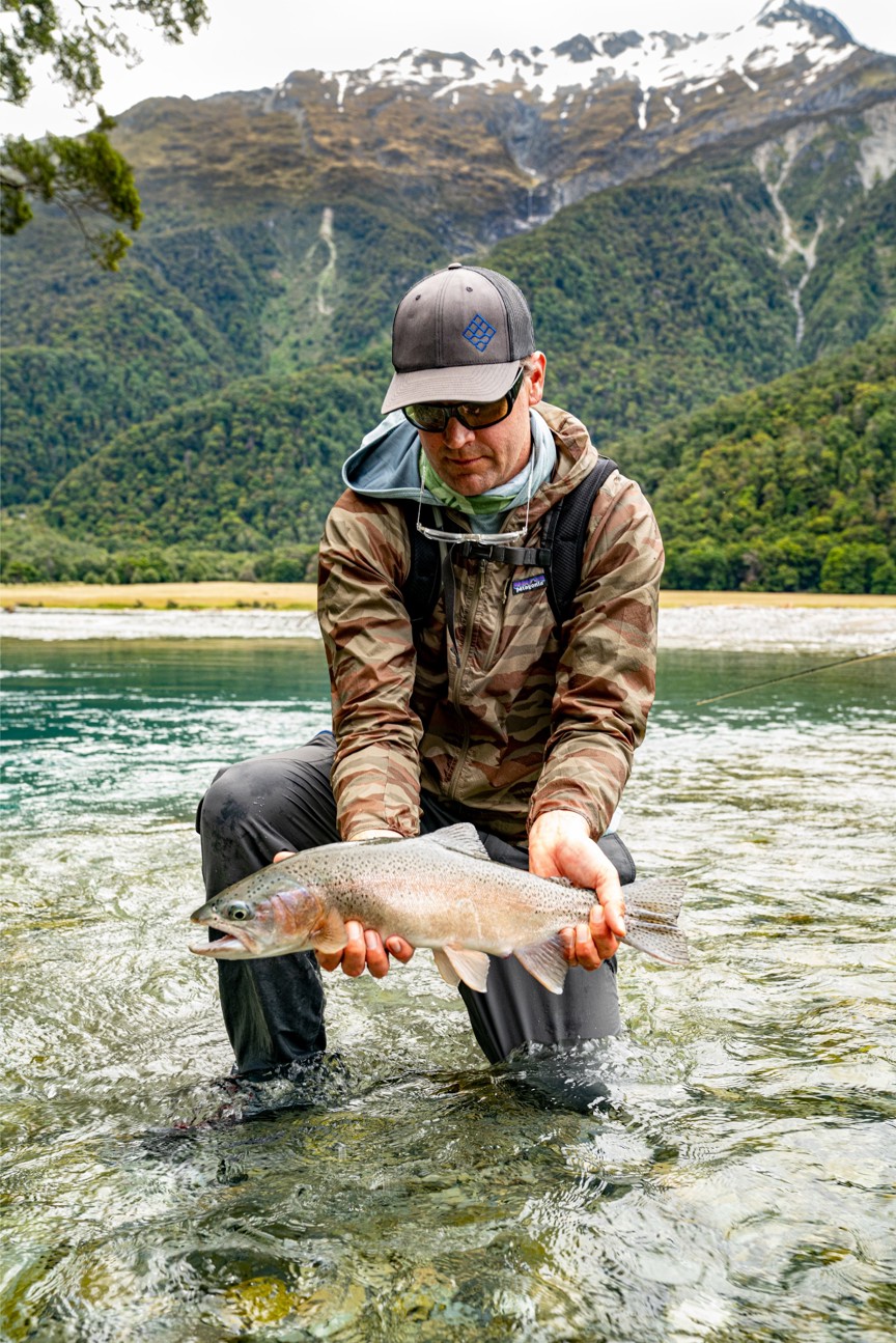 A man holding a caught trout while kneeling in a river.